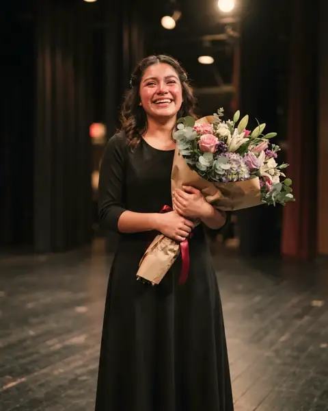 A high school senior in a black concert dress holding a bouquet on a theater stage, tears of joy on her face